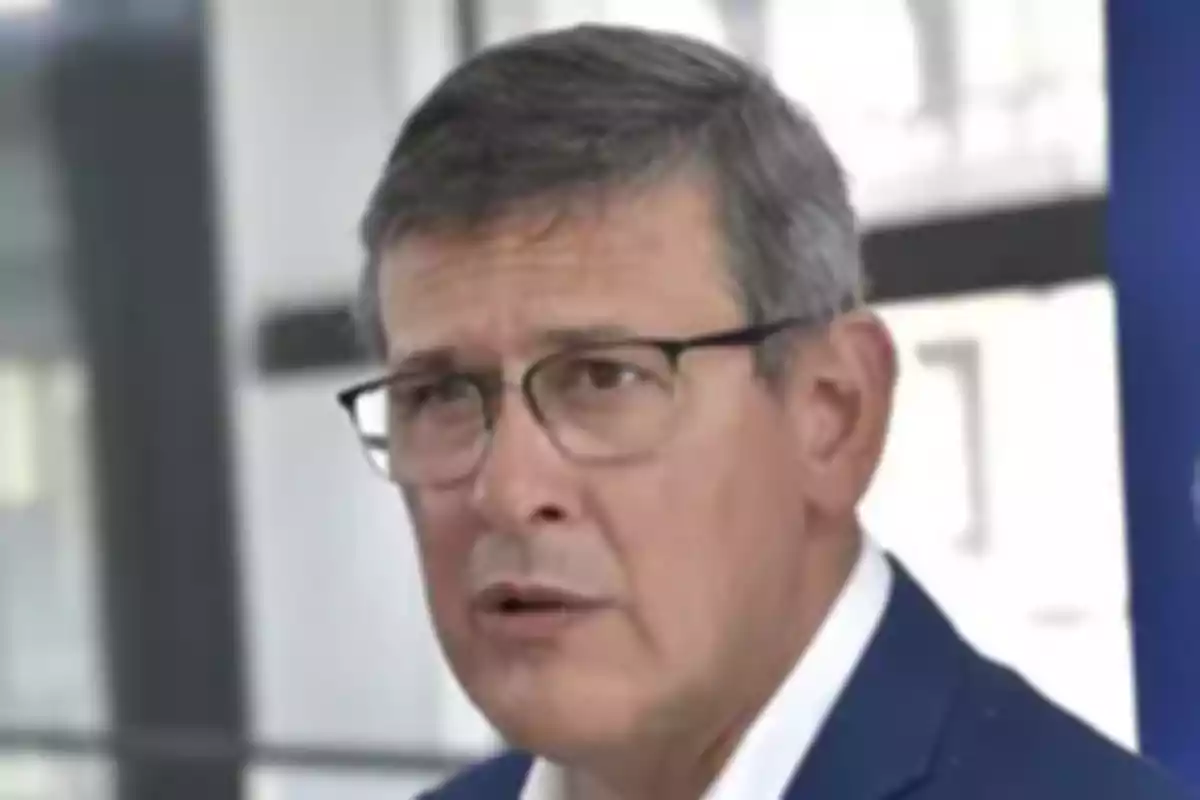Gray-haired man with glasses and a serious expression in a well-lit indoor setting