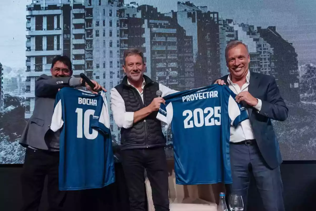 Tres hombres sonrientes posan en un evento sosteniendo camisetas de fútbol azules, una con el nombre Bocha y el número 10, y otra con la palabra Proyectar y el número 2025, con un fondo de edificios urbanos. Tres hombres sonrientes posan en un evento sosteniendo camisetas de fútbol azules, una con el nombre Bocha y el número 10, y otra con la palabra Proyectar y el número 2025, con un fondo de edificios urbanos.