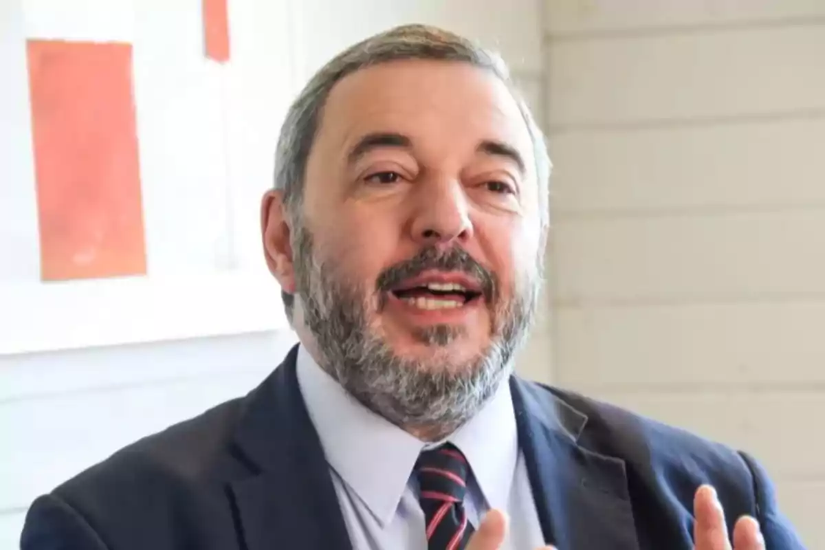 Middle-aged man with a beard and short gray hair, wearing a suit and tie, gesturing while speaking in a well-lit indoor setting