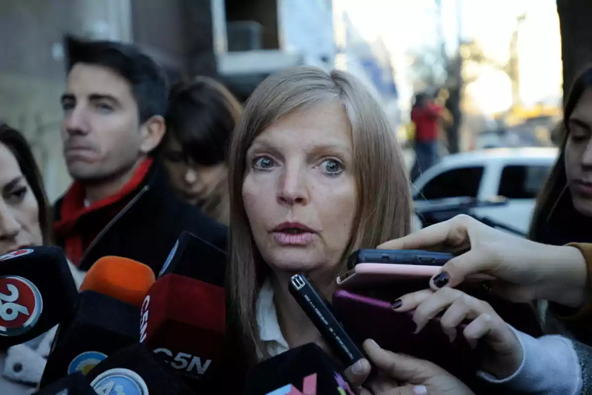 Una mujer rodeada de micrófonos y grabadoras responde preguntas de la prensa en la calle mientras varias personas la observan