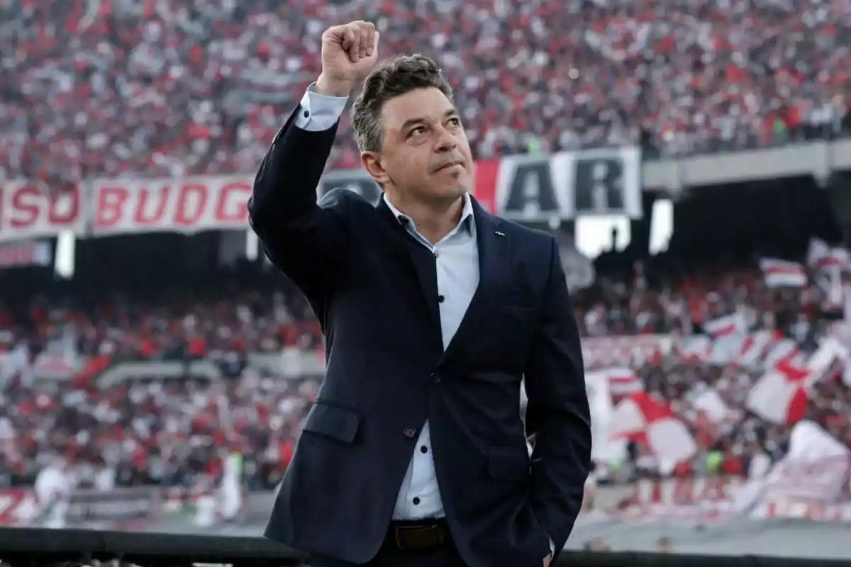 Hombre con traje oscuro levantando el puño en un estadio lleno de aficionados con banderas rojas y blancas