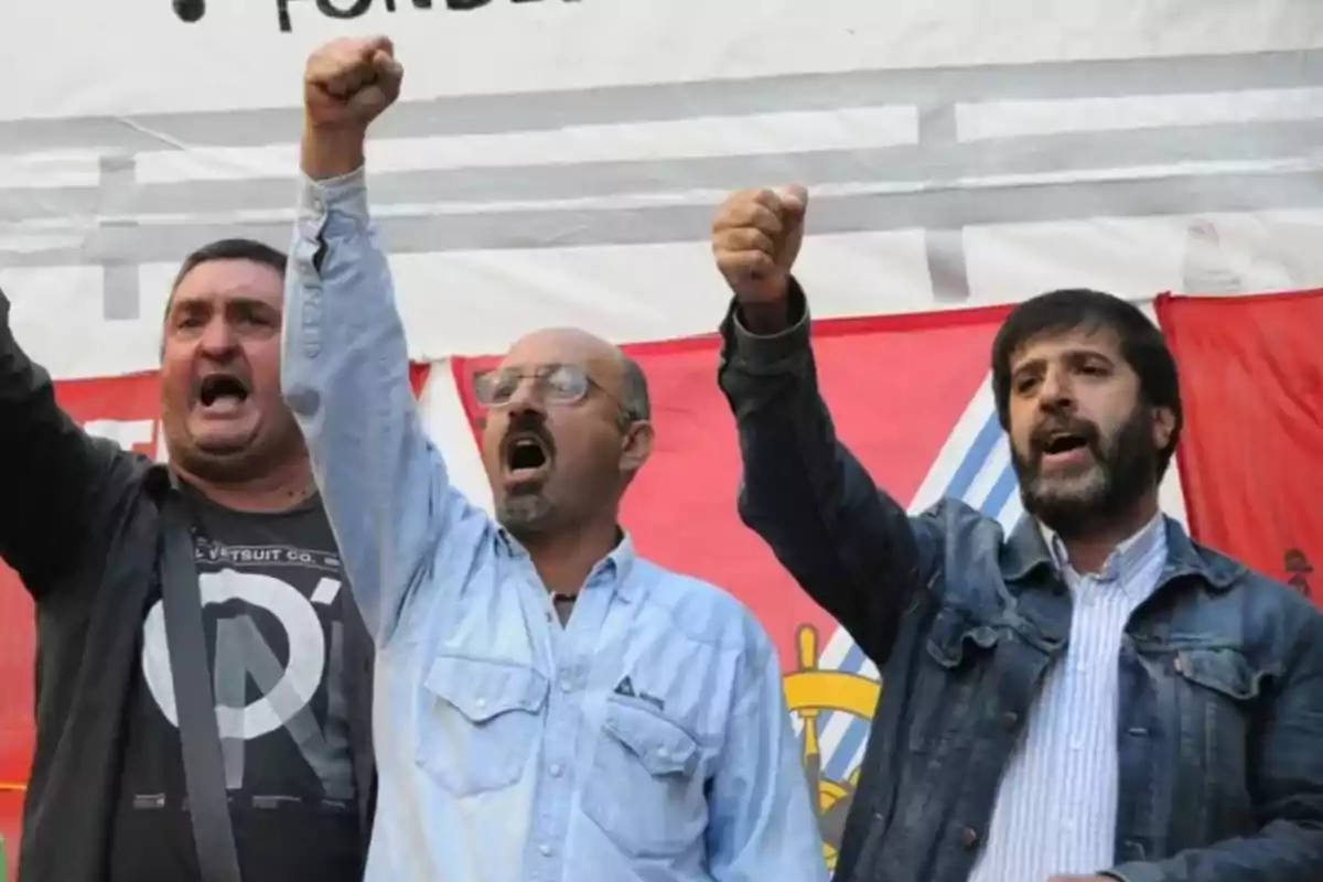 Tres hombres levantan el puño y gritan durante una manifestación frente a una pancarta roja Tres hombres levantan el puño y gritan durante una manifestación frente a una pancarta roja
