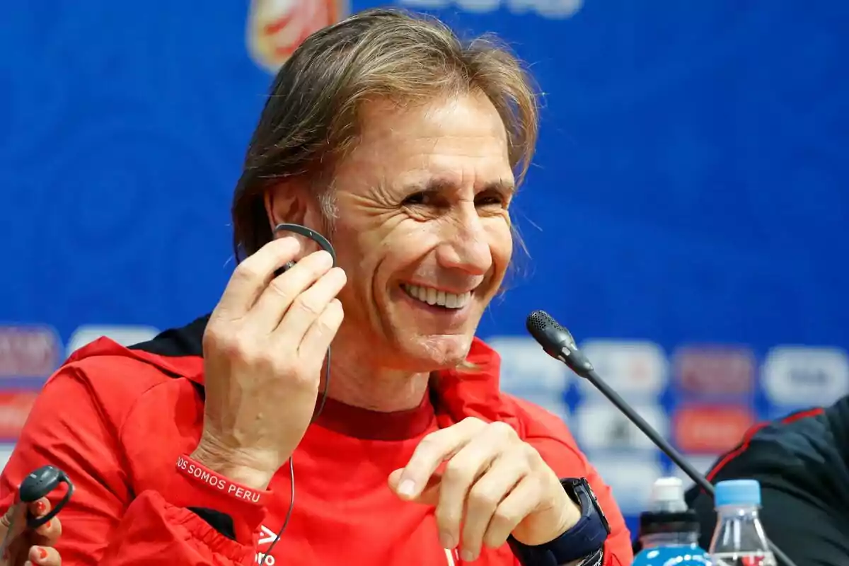 Smiling man in a red jacket at a press conference holding a headphone in front of a microphone