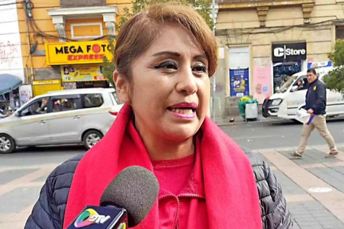 Woman with a red scarf giving an interview on the street in front of a television microphone, with cars and shops in the background