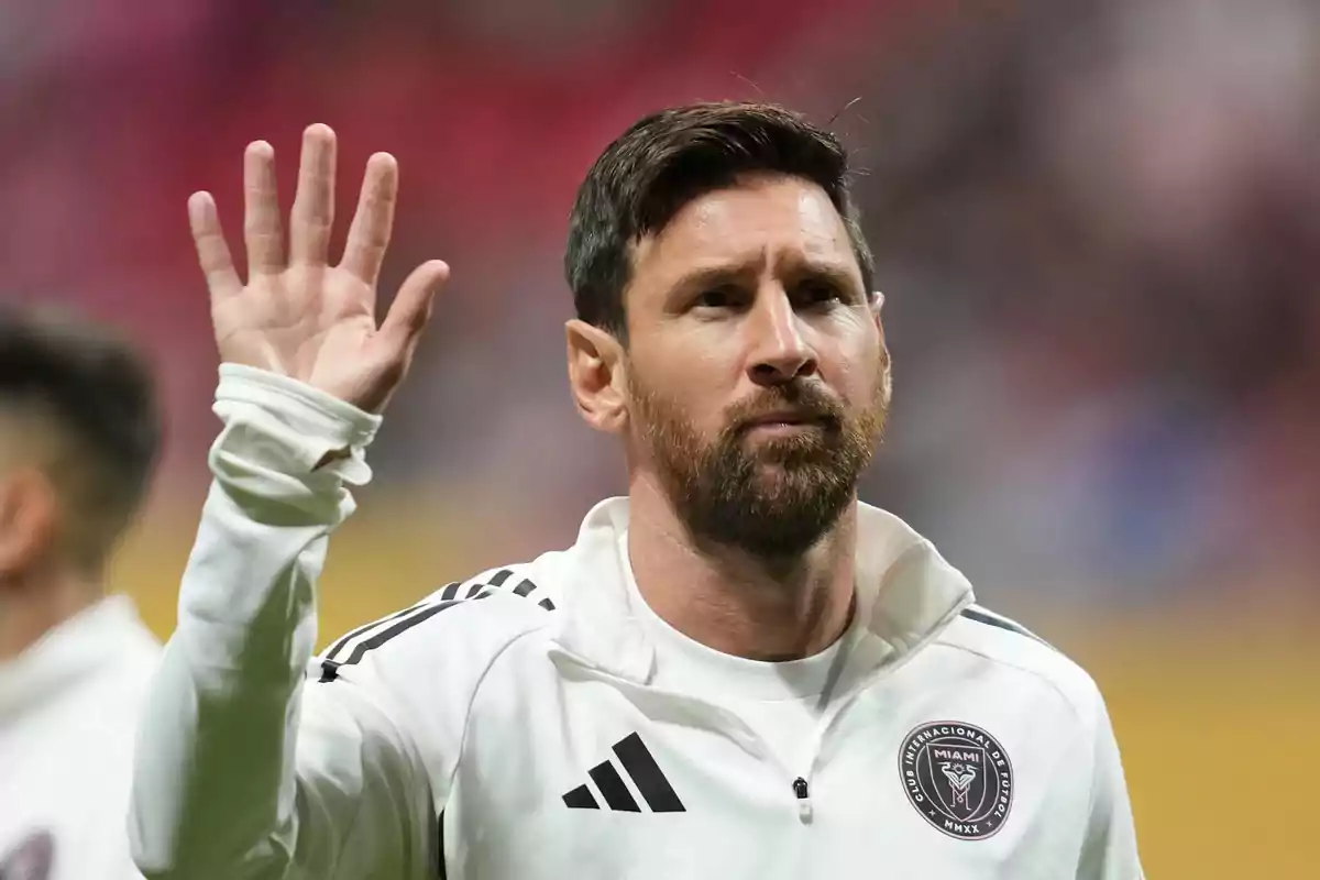 Soccer player with a beard and a white Inter Miami jacket raising his hand in greeting