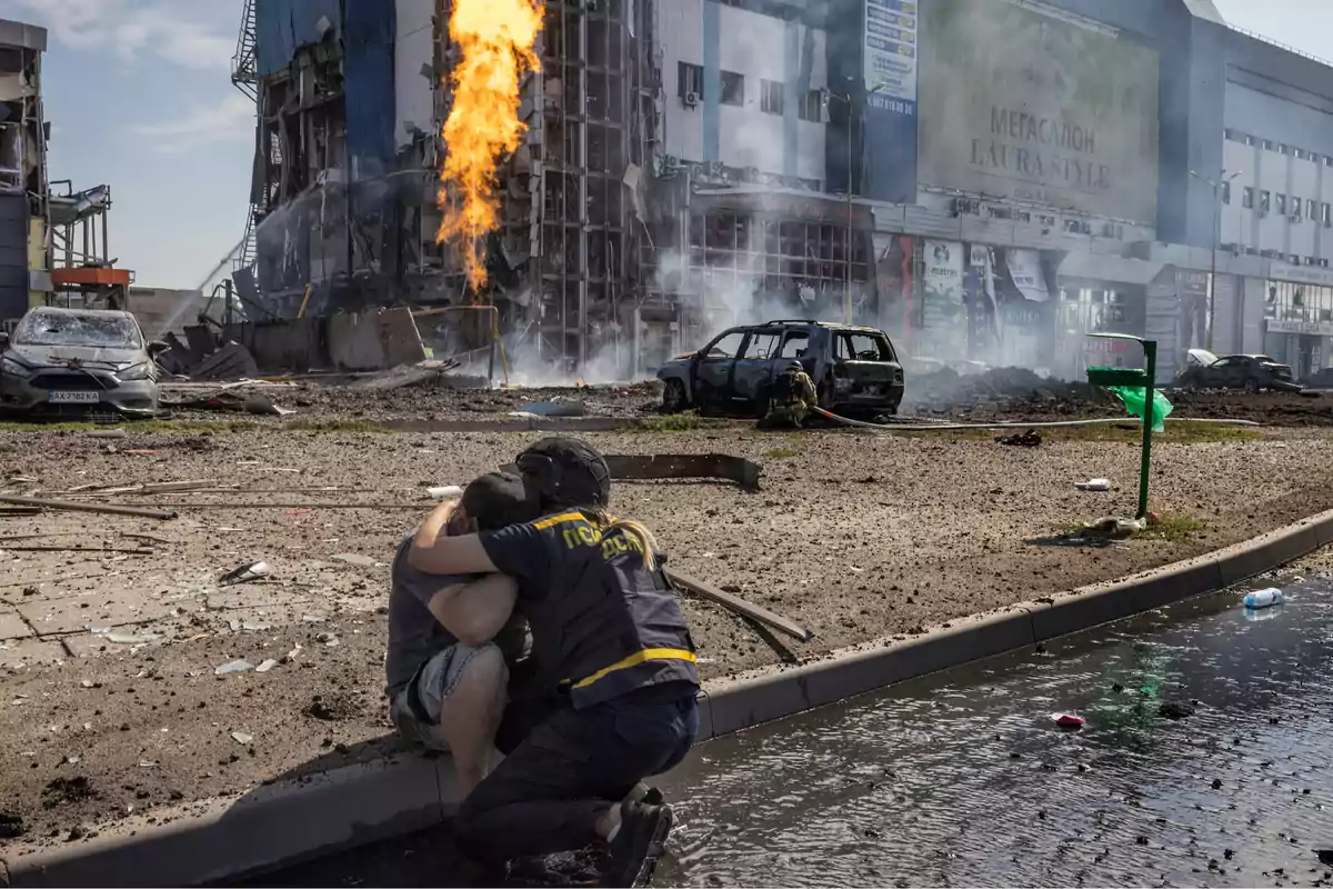 A rescuer comforts a person sitting on the sidewalk while in the background a damaged building, fire, and burned vehicles can be seen after an explosion.