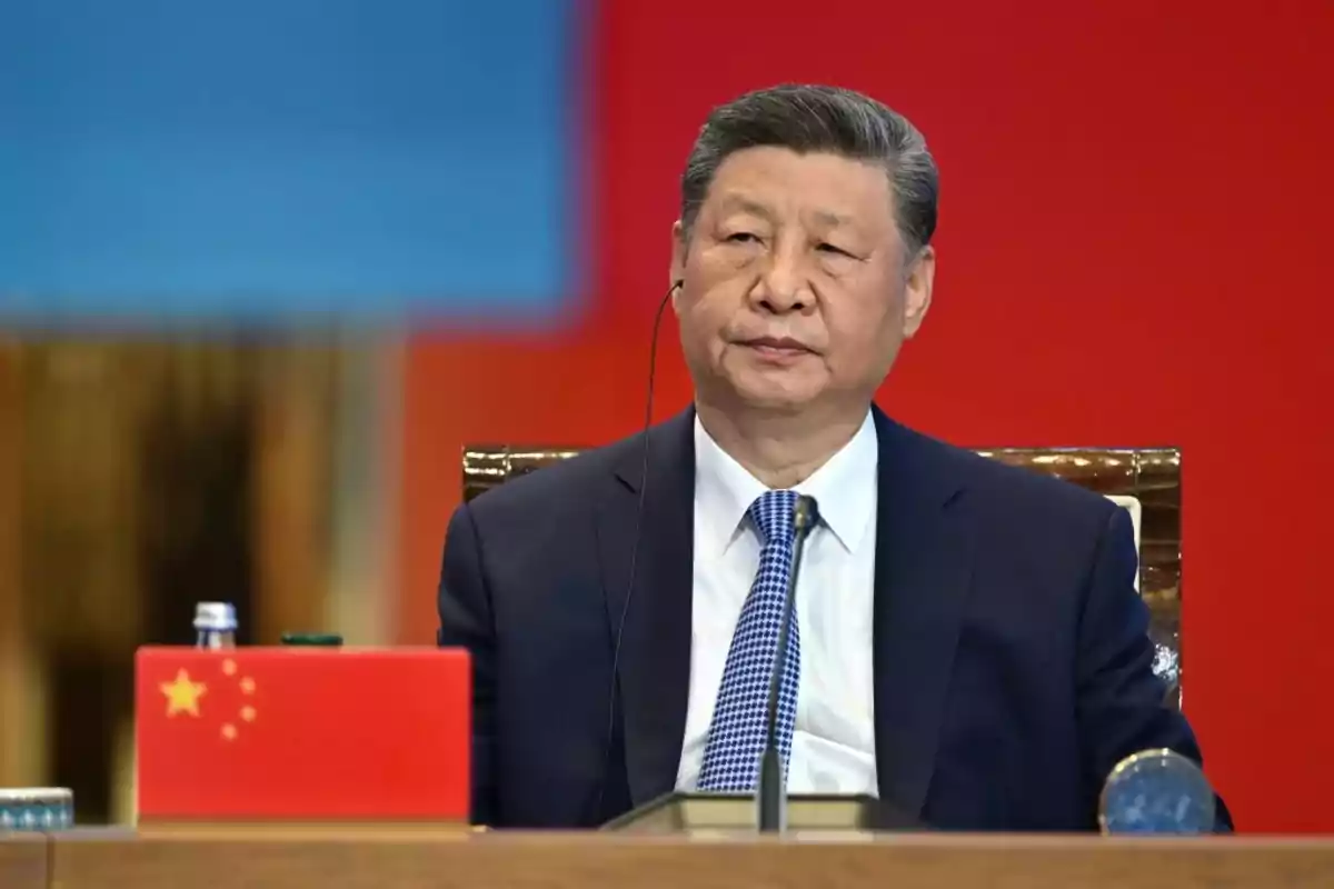 Hombre de traje oscuro y corbata azul sentado frente a una bandera de China en un evento oficial con fondo rojo y azul Hombre de traje oscuro y corbata azul sentado frente a una bandera de China en un evento oficial con fondo rojo y azul