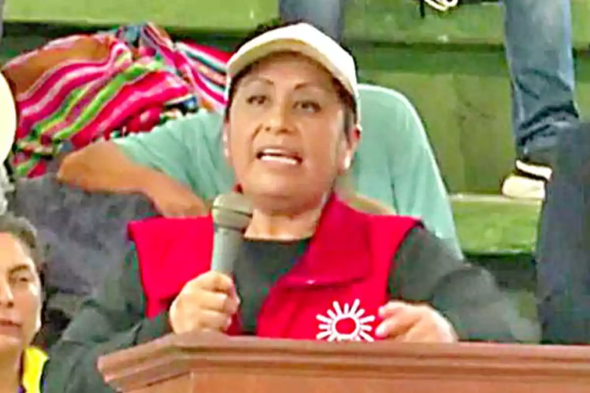 Woman wearing a red vest and beige cap speaking into a microphone behind a podium at a public event