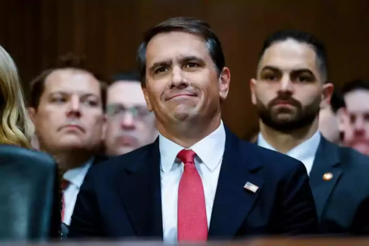 Man in a dark suit and red tie sitting in an audience surrounded by other people in a formal setting Man in a dark suit and red tie sitting in an audience surrounded by other people in a formal setting