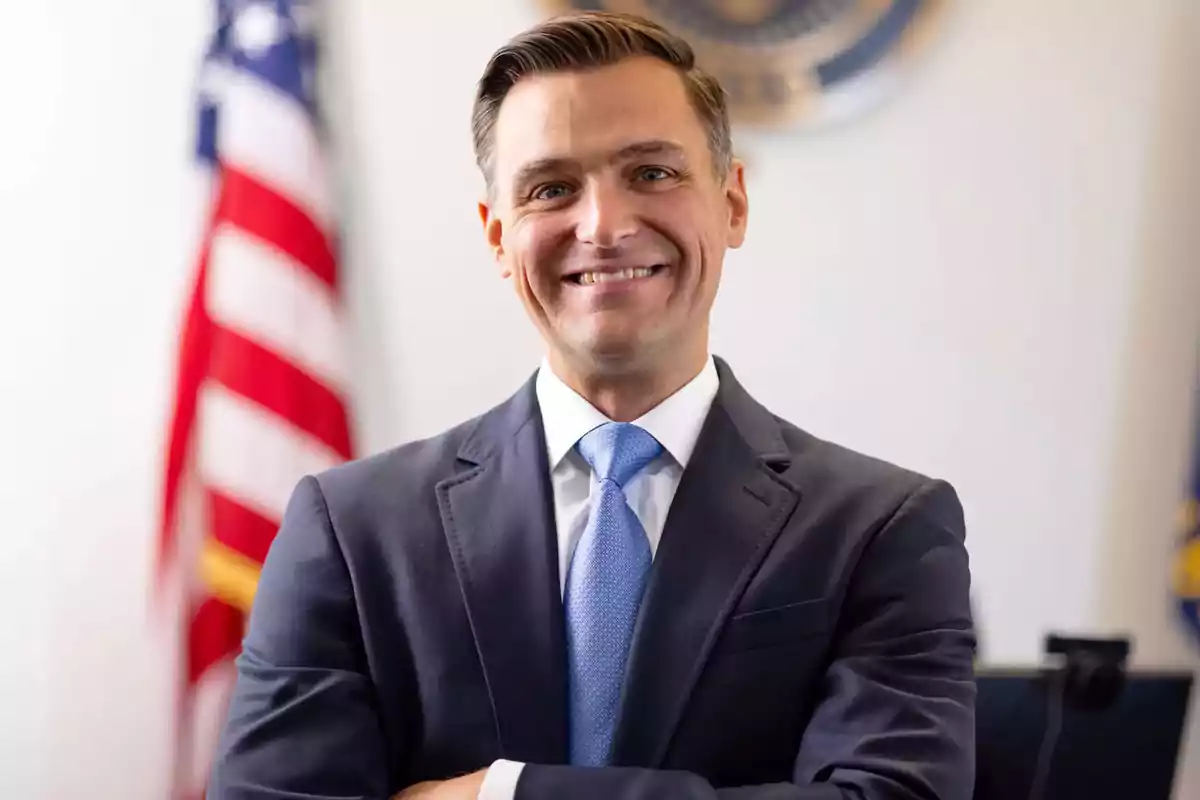 Man in a blue suit and light blue tie smiling with a United States flag in the background Man in a blue suit and light blue tie smiling with a United States flag in the background