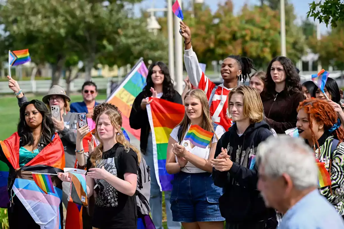 Un grupo de personas jóvenes y adultas se reúne al aire libre sosteniendo banderas del orgullo LGBTQ+ y aplaudiendo durante un evento comunitario en un parque.