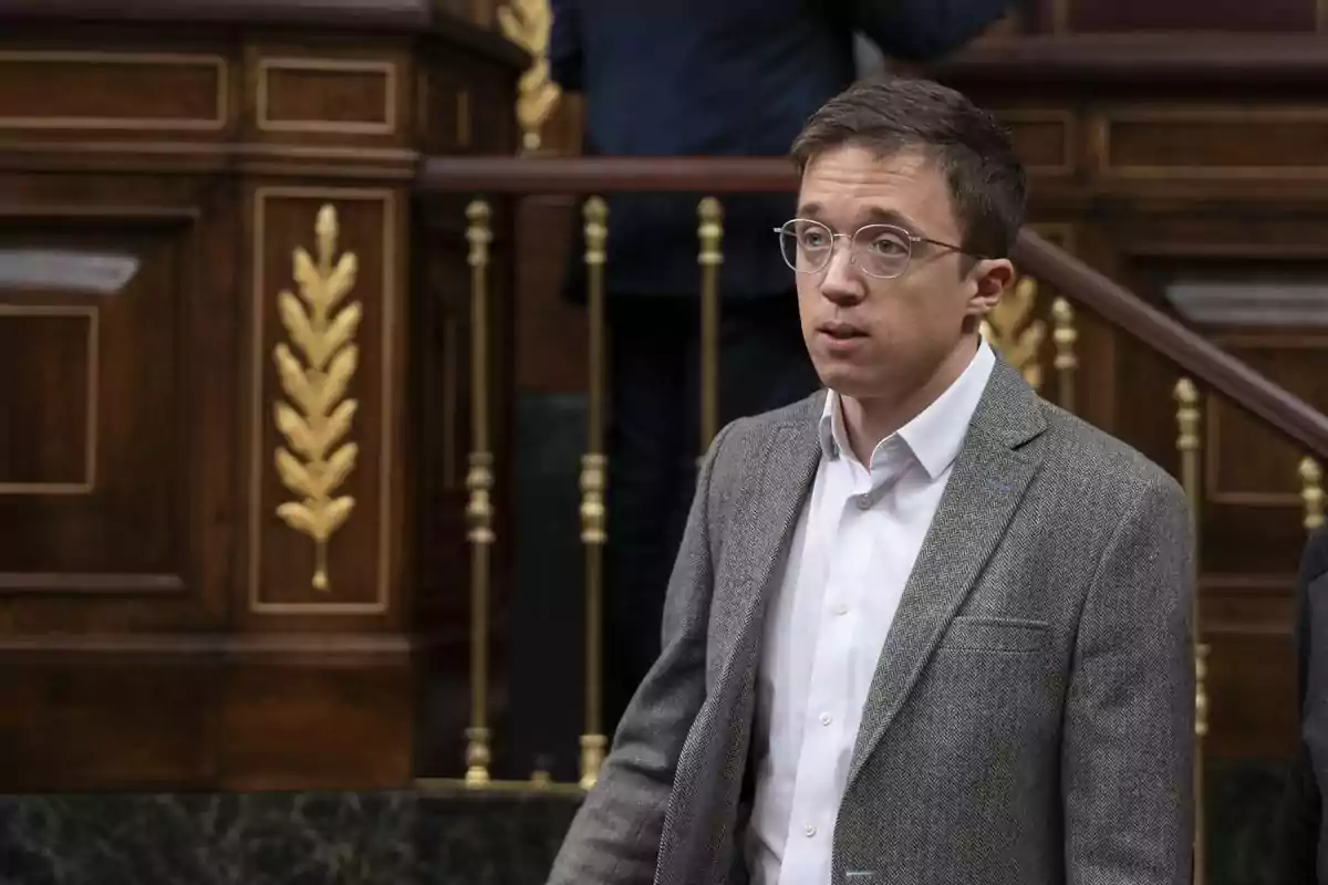 Hombre joven con gafas y chaqueta gris de pie en un entorno parlamentario con detalles dorados en la madera de fondo Hombre joven con gafas y chaqueta gris de pie en un entorno parlamentario con detalles dorados en la madera de fondo
