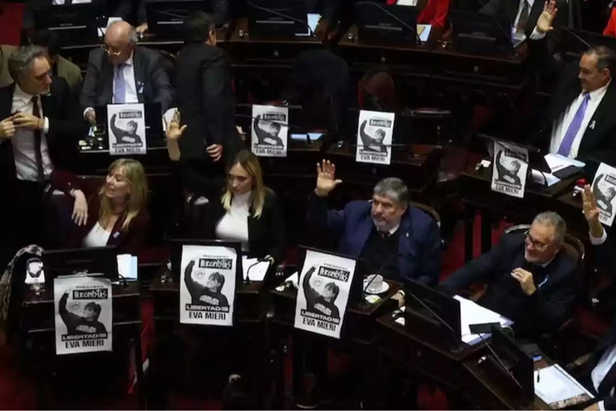 Varios legisladores en un recinto parlamentario levantan la mano mientras en sus escritorios hay carteles con la imagen de una persona y el texto libertad a Eva Mieri