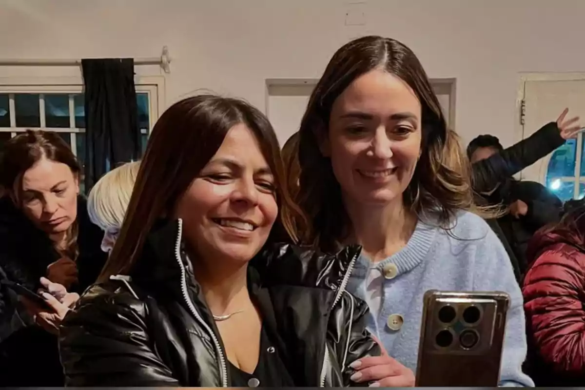 Dos mujeres sonríen mientras se toman una selfie con un teléfono móvil en un ambiente interior con otras personas al fondo Dos mujeres sonríen mientras se toman una selfie con un teléfono móvil en un ambiente interior con otras personas al fondo