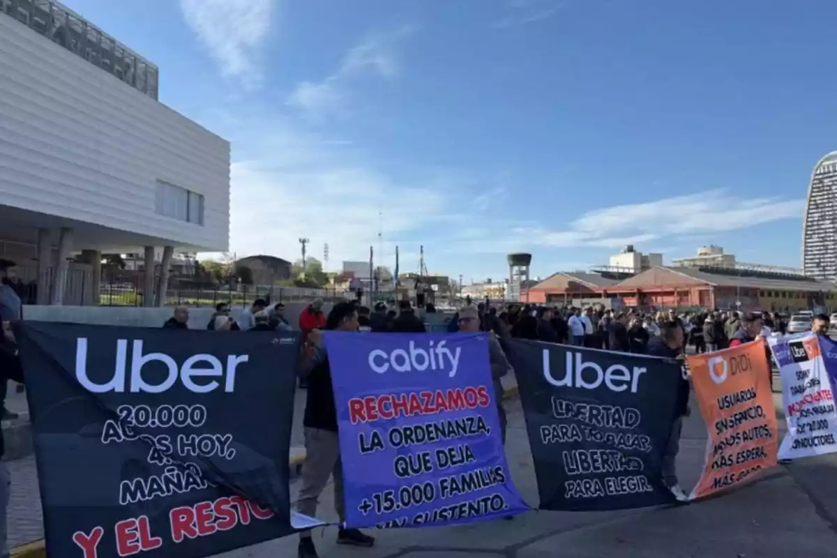 Manifestación de conductores de aplicaciones de transporte como Uber, Cabify y Didi sosteniendo pancartas con mensajes en defensa de su trabajo frente a un edificio moderno y bajo un cielo despejado Manifestación de conductores de aplicaciones de transporte como Uber, Cabify y Didi sosteniendo pancartas con mensajes en defensa de su trabajo frente a un edificio moderno y bajo un cielo despejado