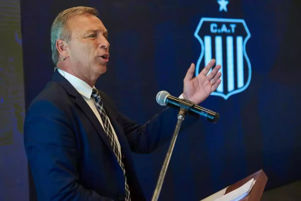 Hombre de traje hablando en un micrófono con el escudo del Club Atlético Talleres de fondo Hombre de traje hablando en un micrófono con el escudo del Club Atlético Talleres de fondo
