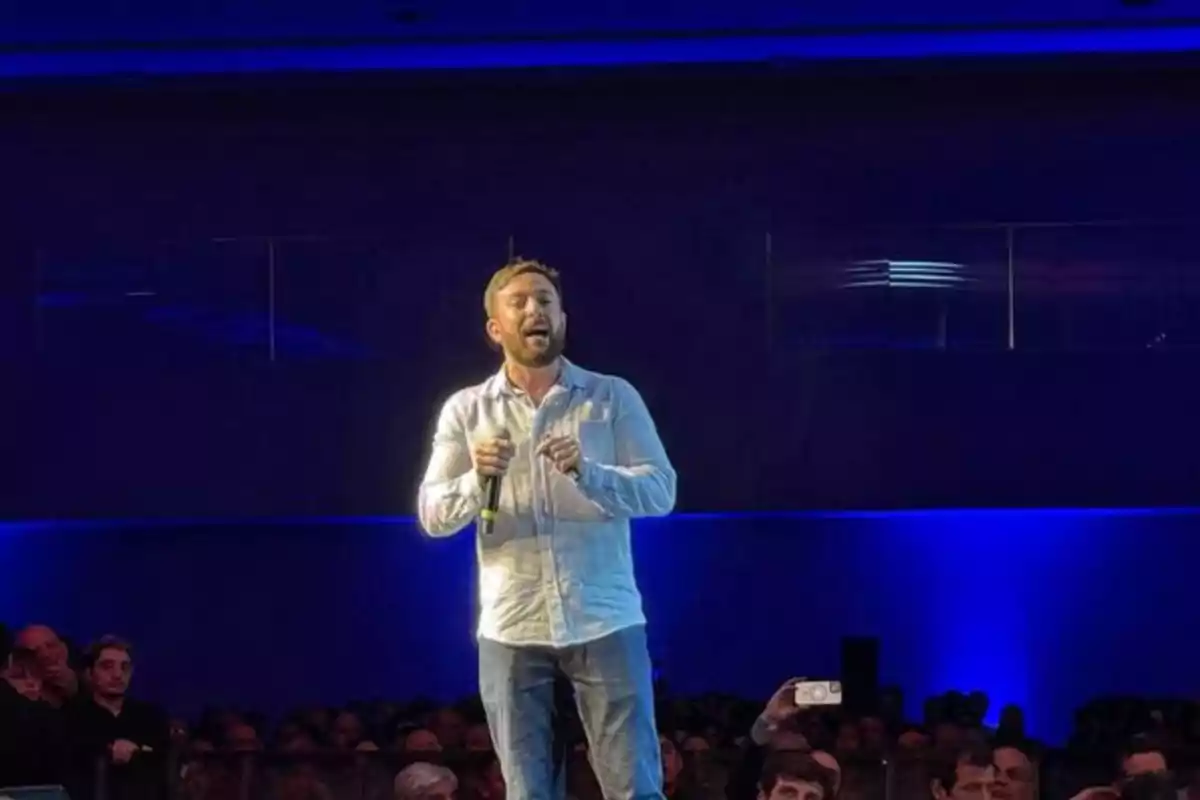 Un hombre con camisa clara y jeans sostiene un micrófono mientras habla en un escenario iluminado con luces azules frente a una audiencia Un hombre con camisa clara y jeans sostiene un micrófono mientras habla en un escenario iluminado con luces azules frente a una audiencia