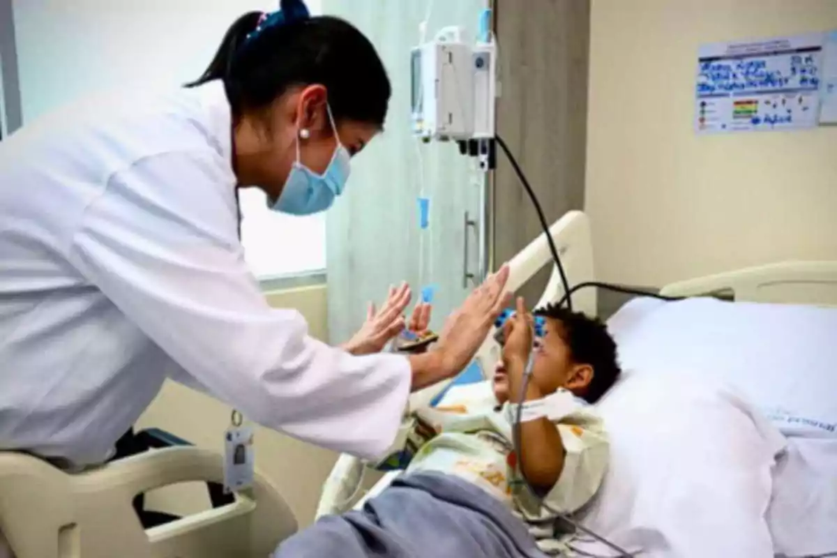 Una doctora con cubrebocas interactúa con un niño hospitalizado que está acostado en una cama y ambos levantan las manos para chocar los cinco