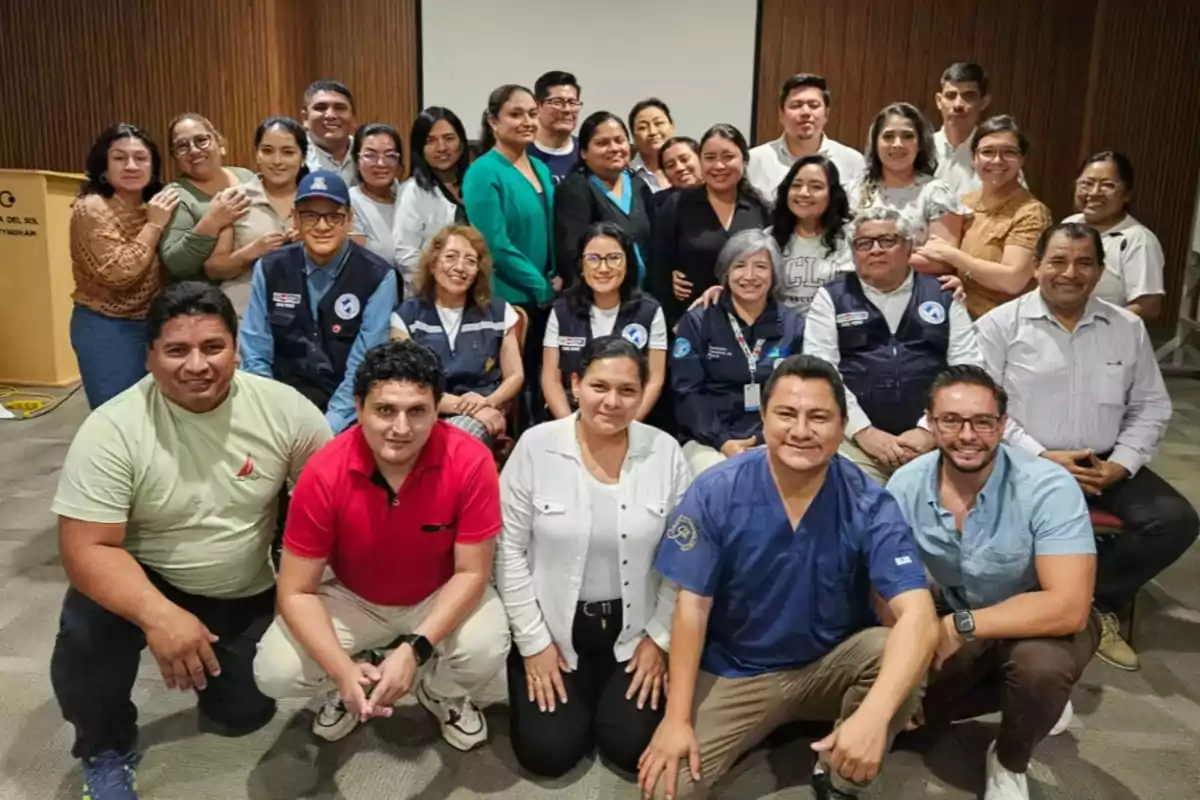 Grupo de personas posando juntas en un salón con paredes de madera y una pantalla al fondo, algunas llevan uniformes y otras ropa casual, todos sonríen hacia la cámara
