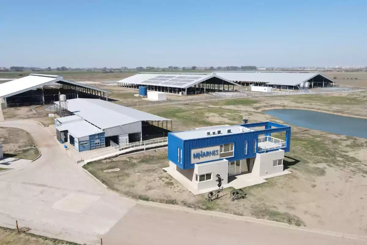 Vista aérea de un complejo agroindustrial con varios galpones de techo blanco, un edificio azul y blanco en primer plano y un pequeño lago artificial al fondo