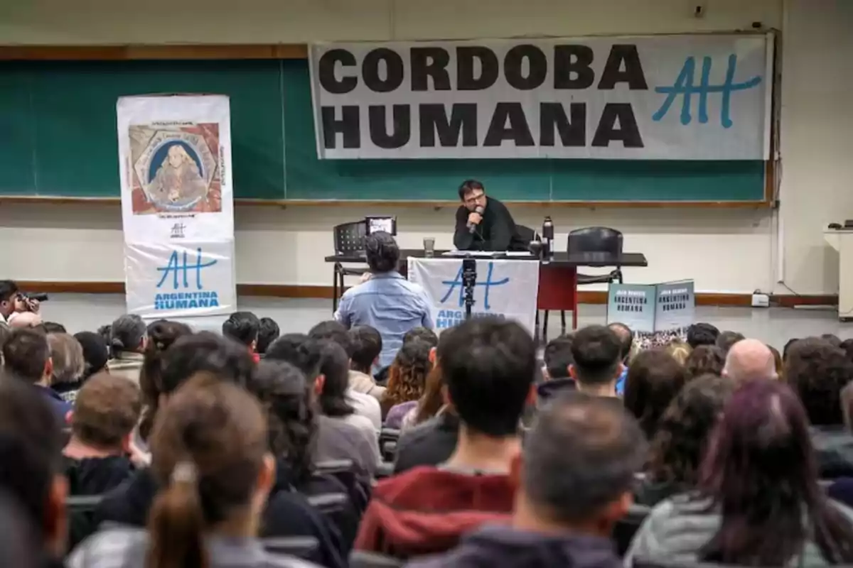 Auditorio lleno de personas escuchando a un orador sentado en una mesa frente a un pizarrón, con carteles que dicen Córdoba Humana y Argentina Humana.