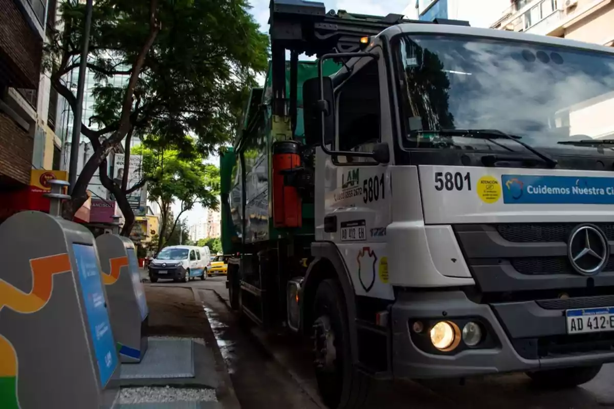 Camión de basura realizando la recolección de residuos en una calle urbana junto a contenedores de reciclaje y árboles