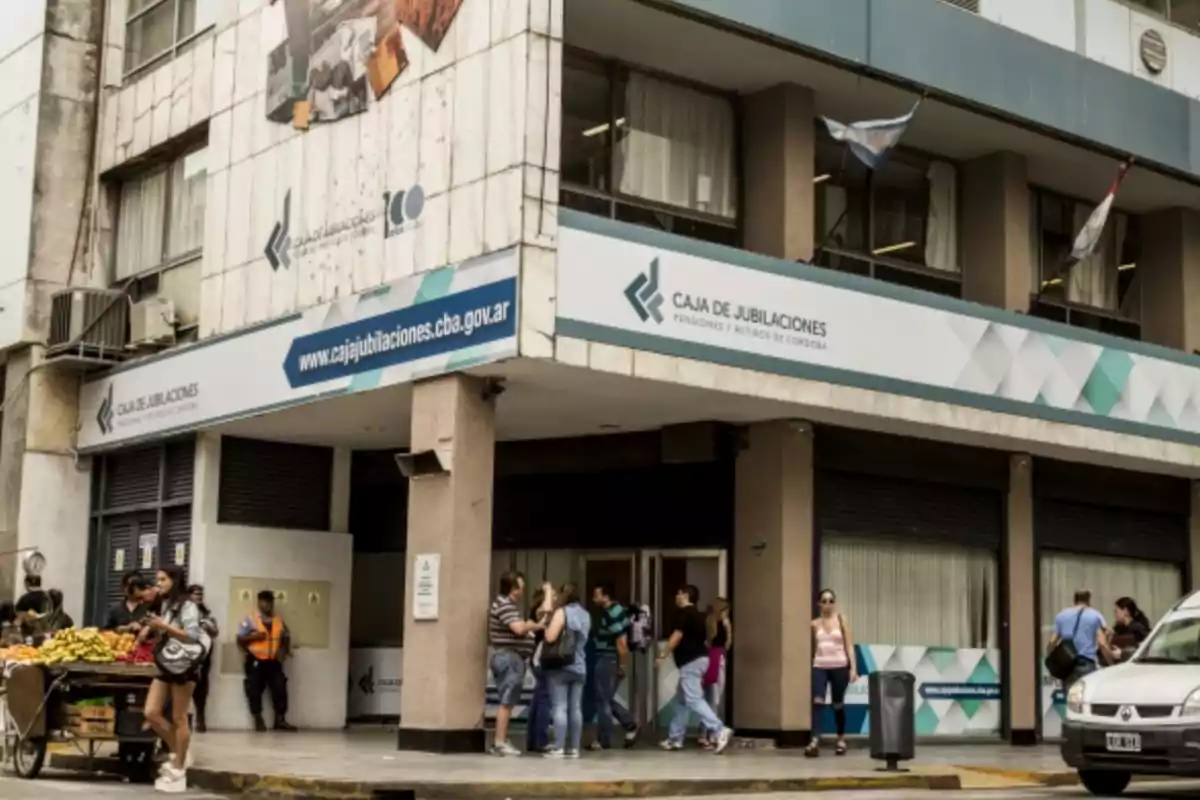 Personas entrando y saliendo de un edificio de la Caja de Jubilaciones de Córdoba mientras una mujer compra frutas en un puesto callejero Personas entrando y saliendo de un edificio de la Caja de Jubilaciones de Córdoba mientras una mujer compra frutas en un puesto callejero