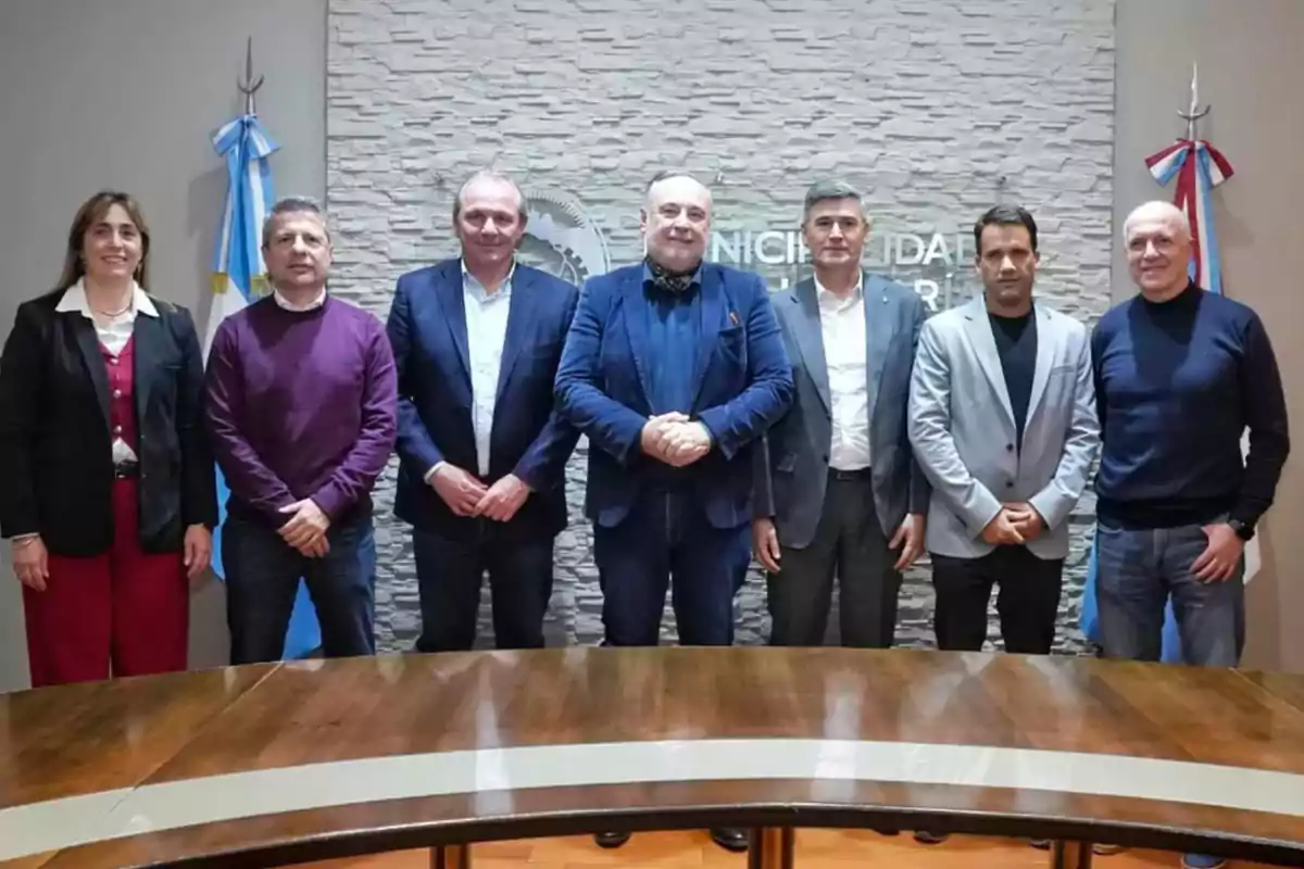 Seven people stand in front of a wooden table in a room with Argentine flags and a background wall featuring an institutional logo.