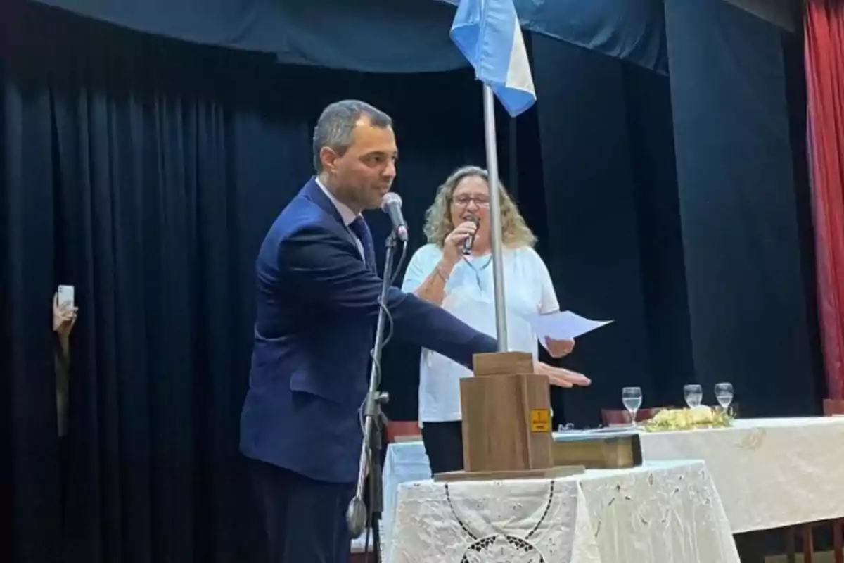 Un hombre de traje participa en un acto formal junto a una mujer que sostiene un micrófono y papeles, ambos están en un escenario con una bandera celeste y blanca y una mesa decorada al frente.
