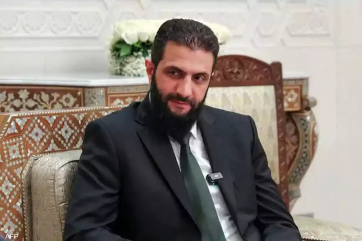 Hombre de barba oscura y cabello corto vestido con traje negro y corbata verde sentado en un sillón decorado con patrones geométricos y fondo elegante
