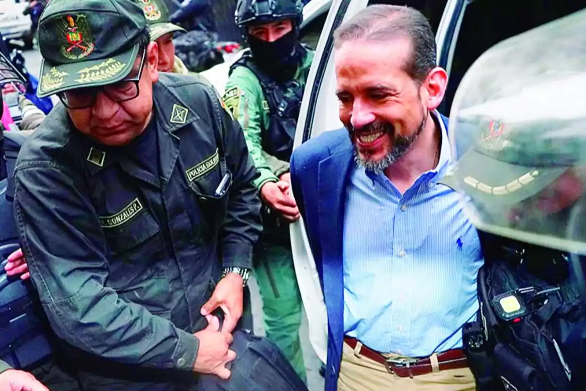 Un hombre con barba y saco azul es escoltado por varios policías uniformados mientras sale de un vehículo
