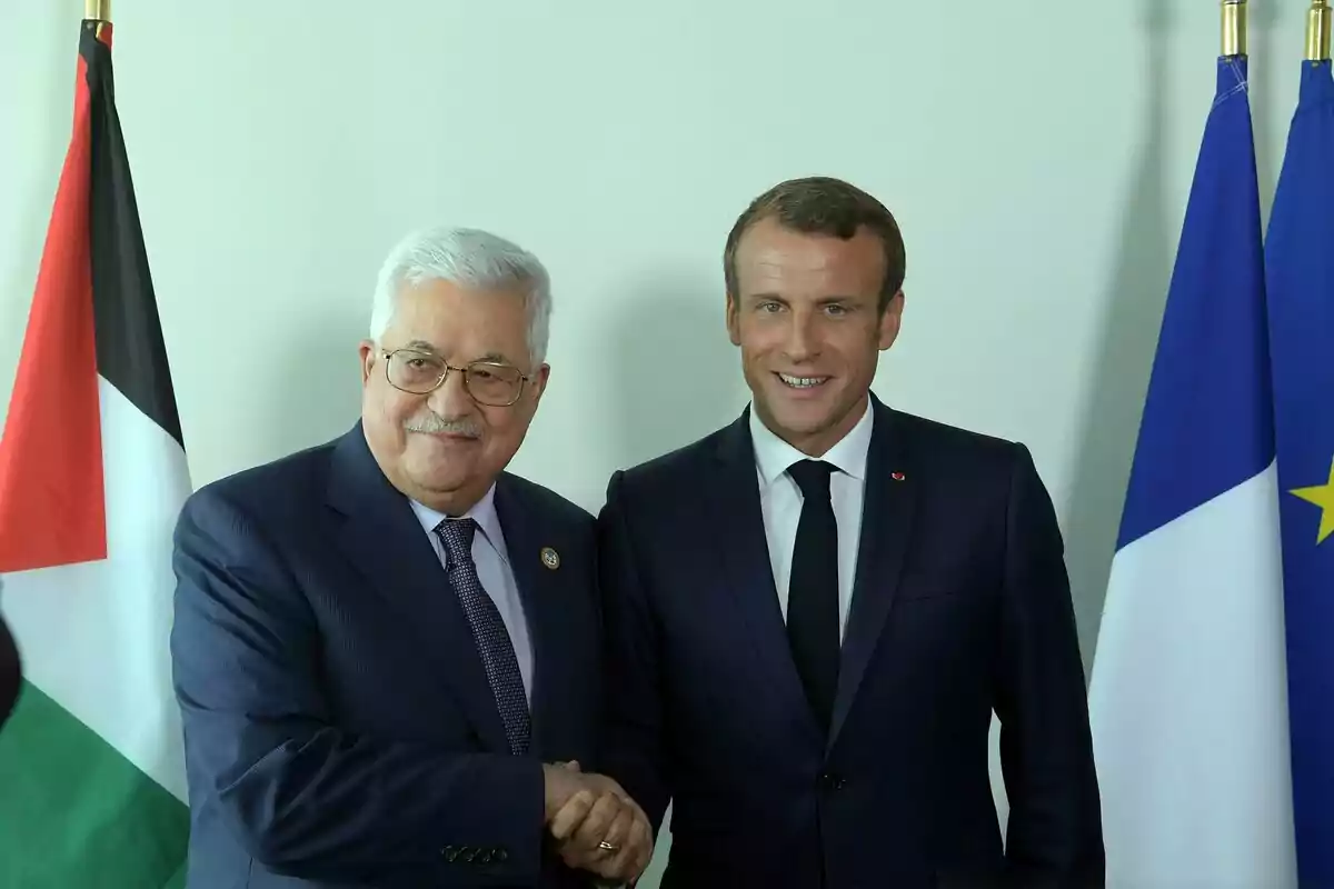 Two men in suits shake hands and smile in front of the flags of Palestine and France.