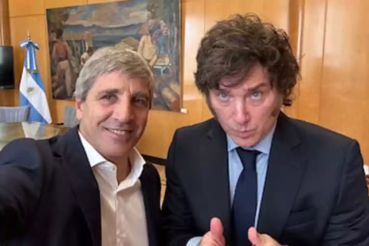 Two men in suits pose for a selfie in an office with an Argentine flag and a painting on the wall in the background.