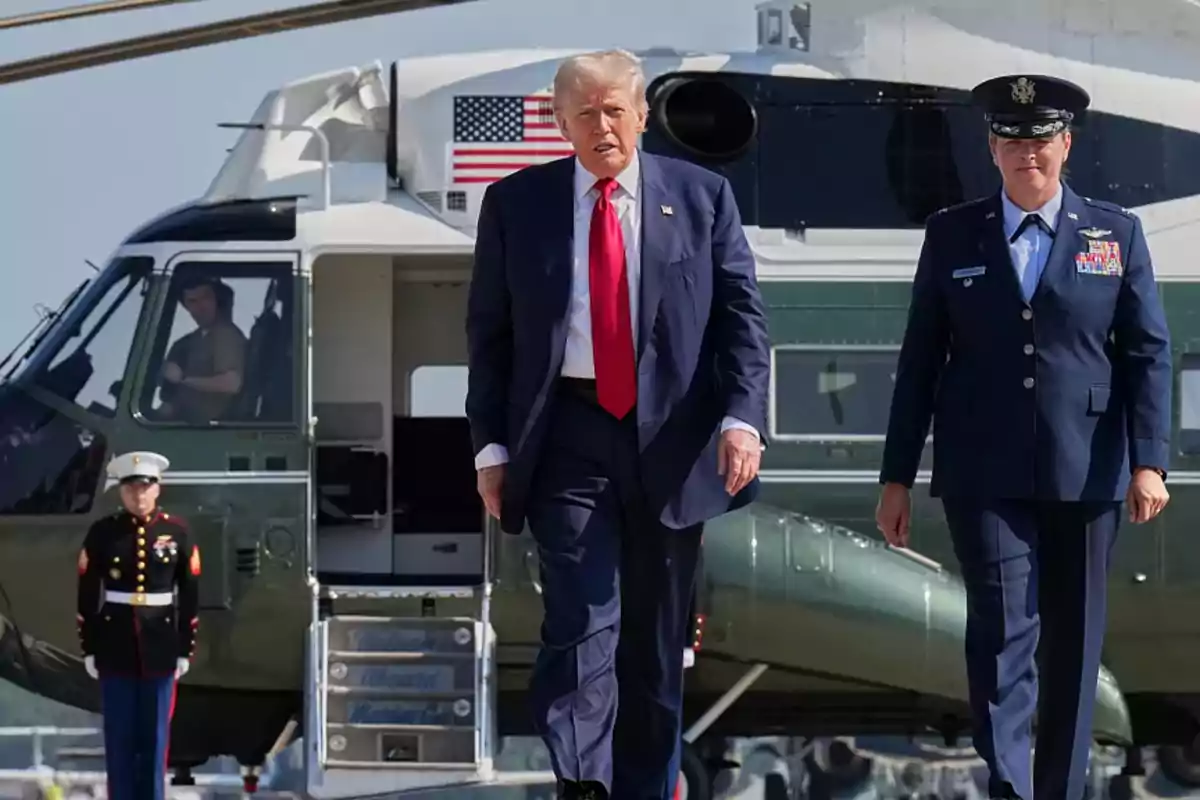 A man in a blue suit and red tie walks alongside a military officer in front of a helicopter with the United States flag visible in the background. A man in a blue suit and red tie walks alongside a military officer in front of a helicopter with the United States flag visible in the background.