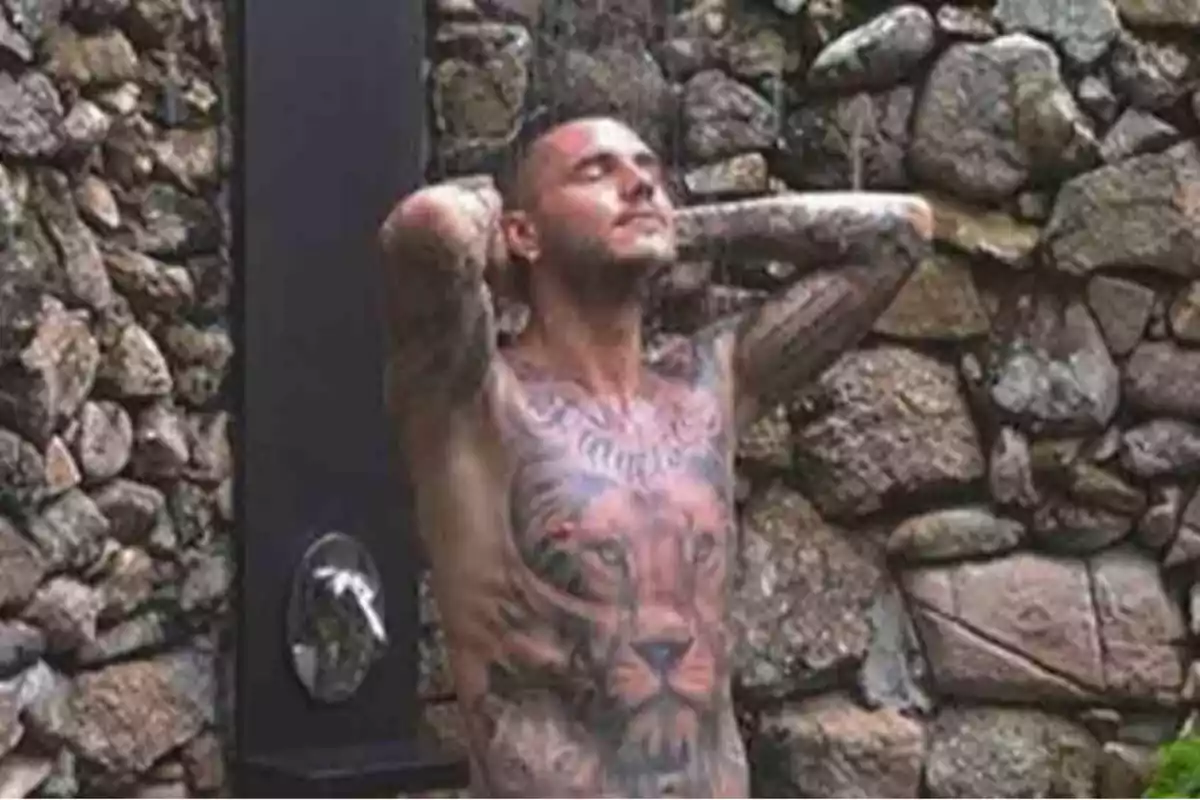 Hombre tatuado disfrutando de una ducha al aire libre frente a una pared de piedras