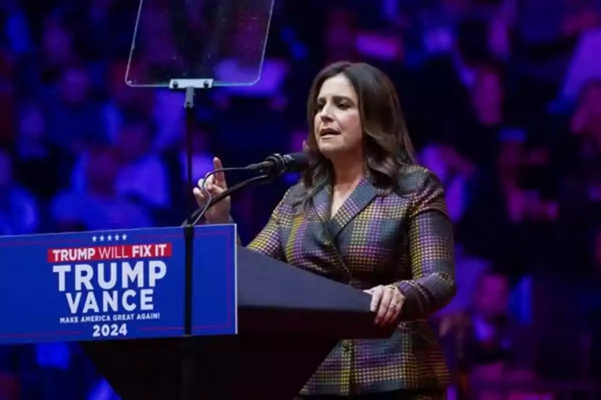Mujer hablando en un mitin político frente a un podio con un cartel de campaña de Trump Vance 2024 y fondo iluminado en tonos morados y azules Mujer hablando en un mitin político frente a un podio con un cartel de campaña de Trump Vance 2024 y fondo iluminado en tonos morados y azules
