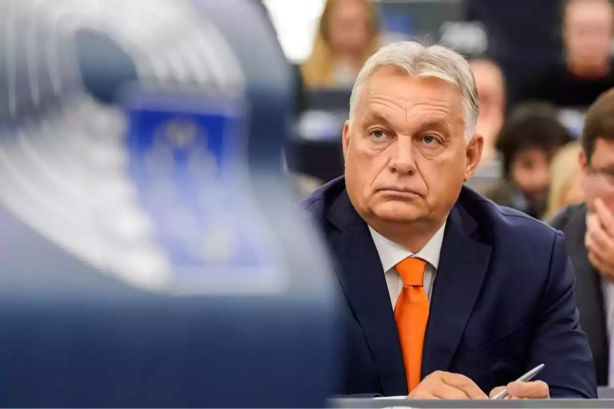 Gray-haired man in a blue suit and orange tie sitting in a conference room with people in the background Gray-haired man in a blue suit and orange tie sitting in a conference room with people in the background