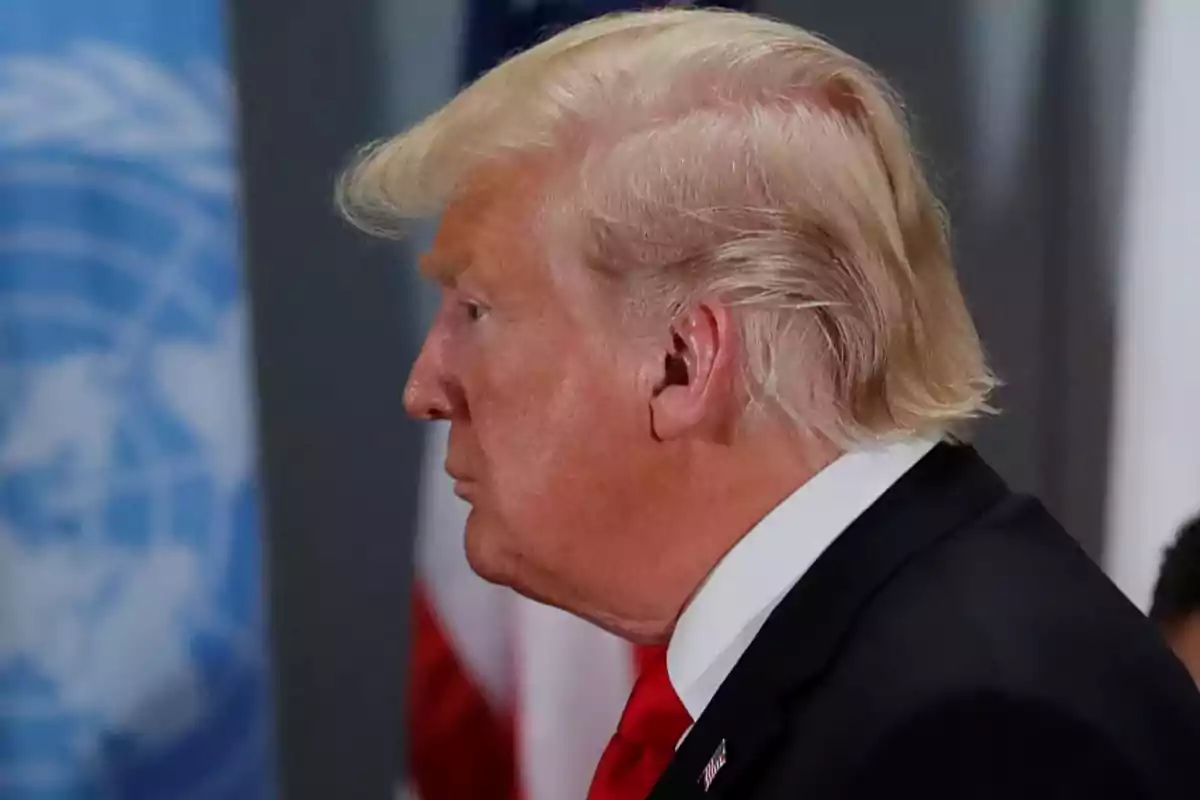 Man in profile with blond hair and a dark suit in front of a blurred UN flag Man in profile with blond hair and a dark suit in front of a blurred UN flag