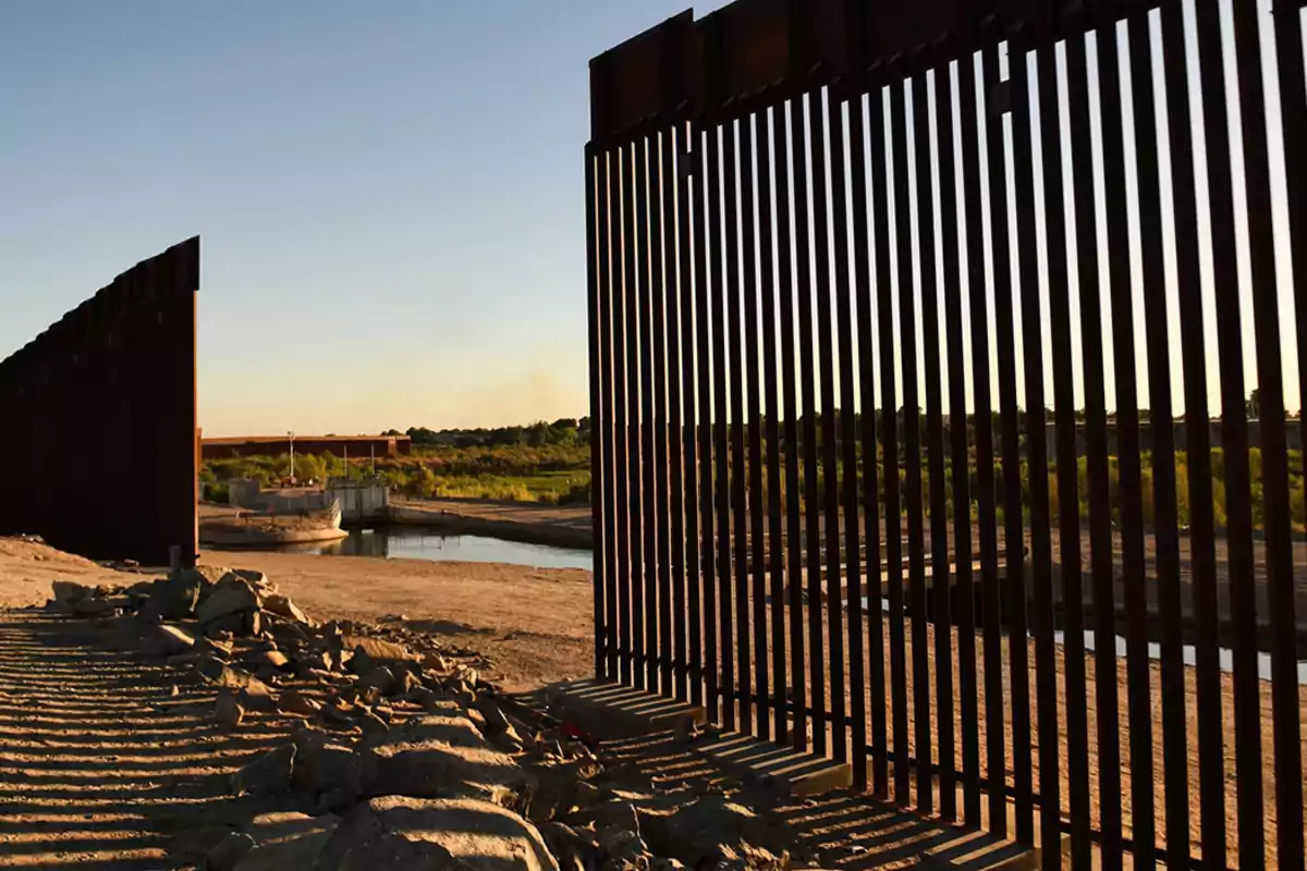 A tall metal fence is seen next to a river and rocky terrain at sunset, with vegetation in the background. A tall metal fence is seen next to a river and rocky terrain at sunset, with vegetation in the background.
