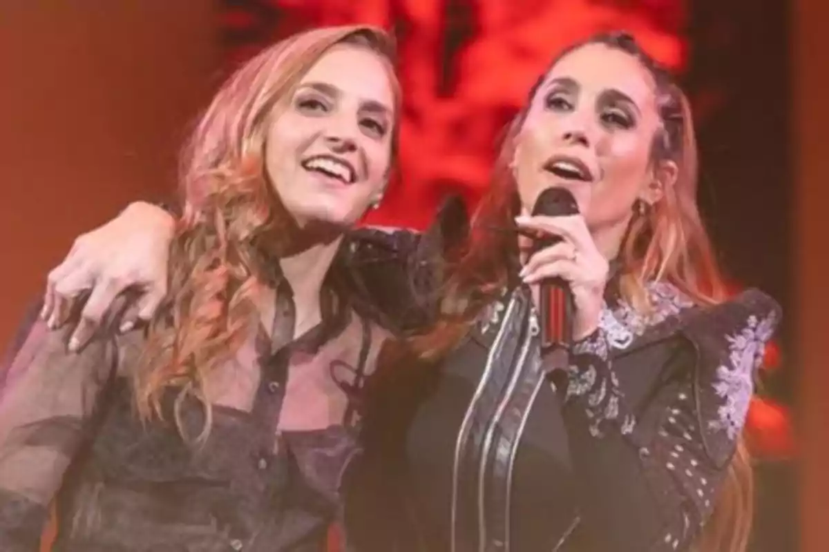 Dos mujeres cantan juntas en el escenario mientras una de ellas sostiene un micrófono y la otra sonríe abrazándola Dos mujeres cantan juntas en el escenario mientras una de ellas sostiene un micrófono y la otra sonríe abrazándola