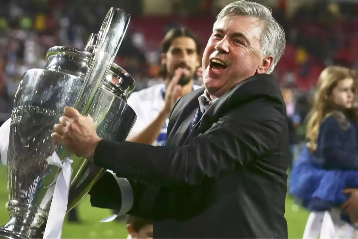 Smiling gray-haired man holds a large silver trophy in a stadium full of people celebrating a sports victory Smiling gray-haired man holds a large silver trophy in a stadium full of people celebrating a sports victory