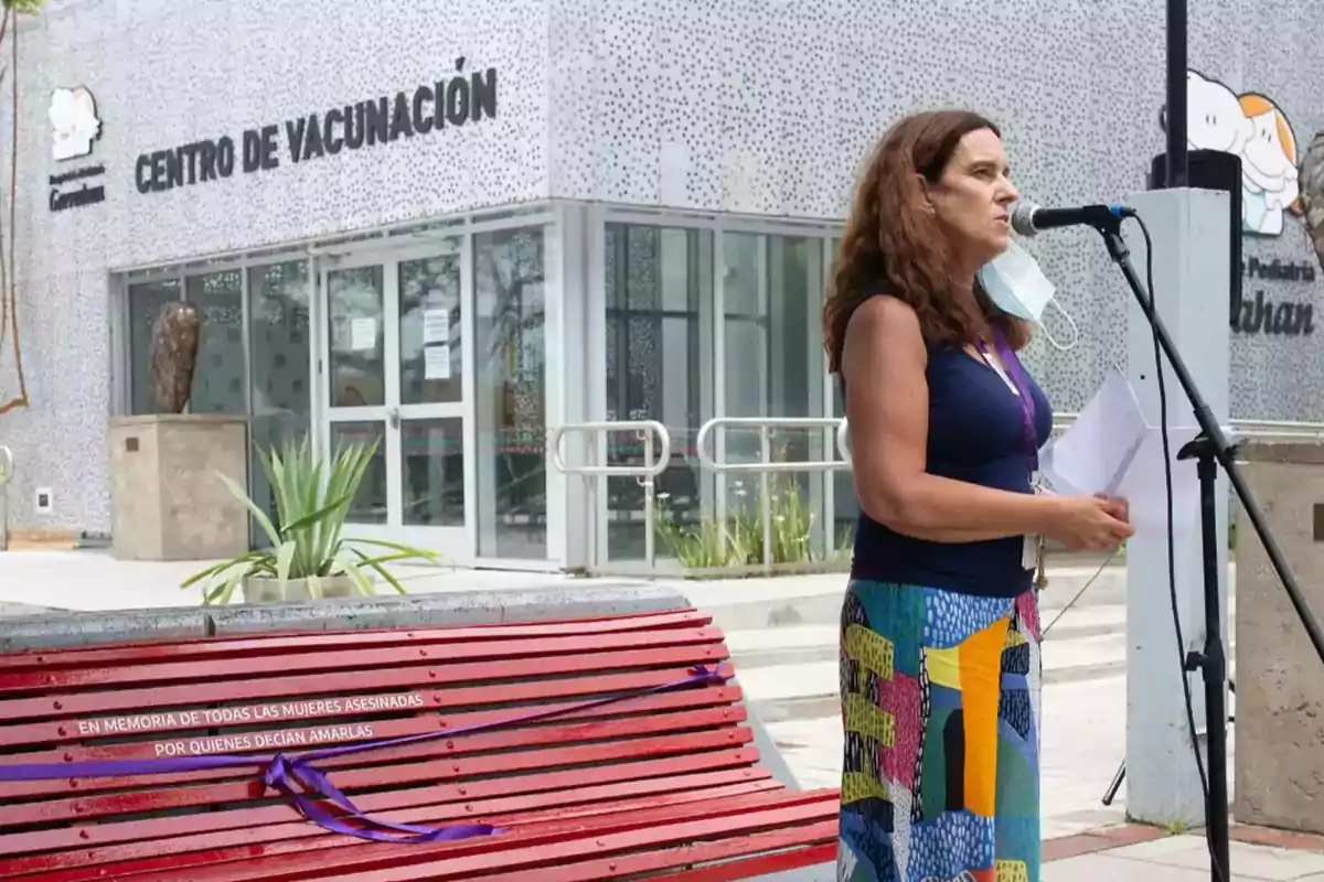Mujer hablando frente a un micrófono afuera de un centro de vacunación con un banco rojo en primer plano que tiene un listón morado y un mensaje en memoria de mujeres asesinadas Mujer hablando frente a un micrófono afuera de un centro de vacunación con un banco rojo en primer plano que tiene un listón morado y un mensaje en memoria de mujeres asesinadas
