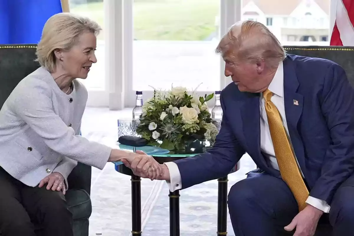 Dos personas elegantemente vestidas se dan la mano sentadas frente a una mesa con un arreglo floral