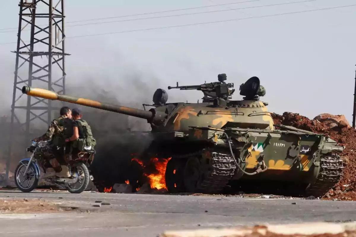 Dos hombres en motocicleta pasan junto a un tanque militar en llamas en una zona de conflicto