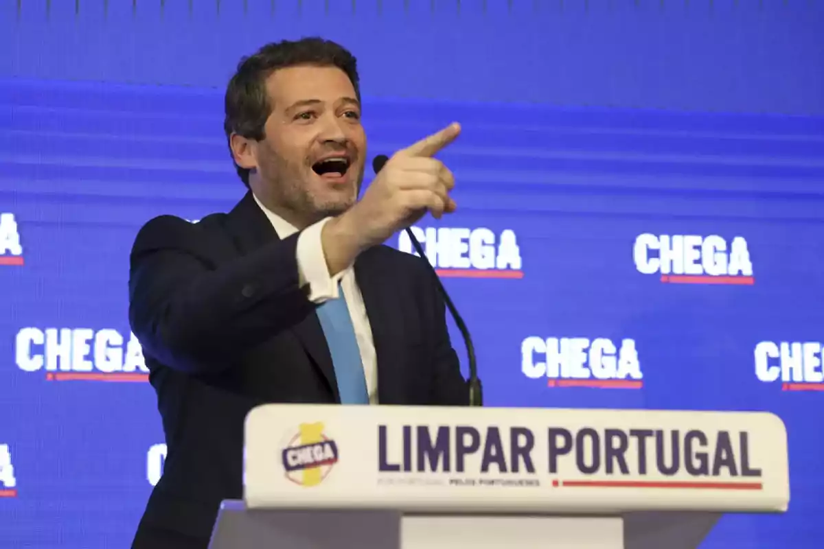 Man in a suit speaking and pointing during a speech at a podium with the text Limpar Portugal and a blue background with the word Chega repeated