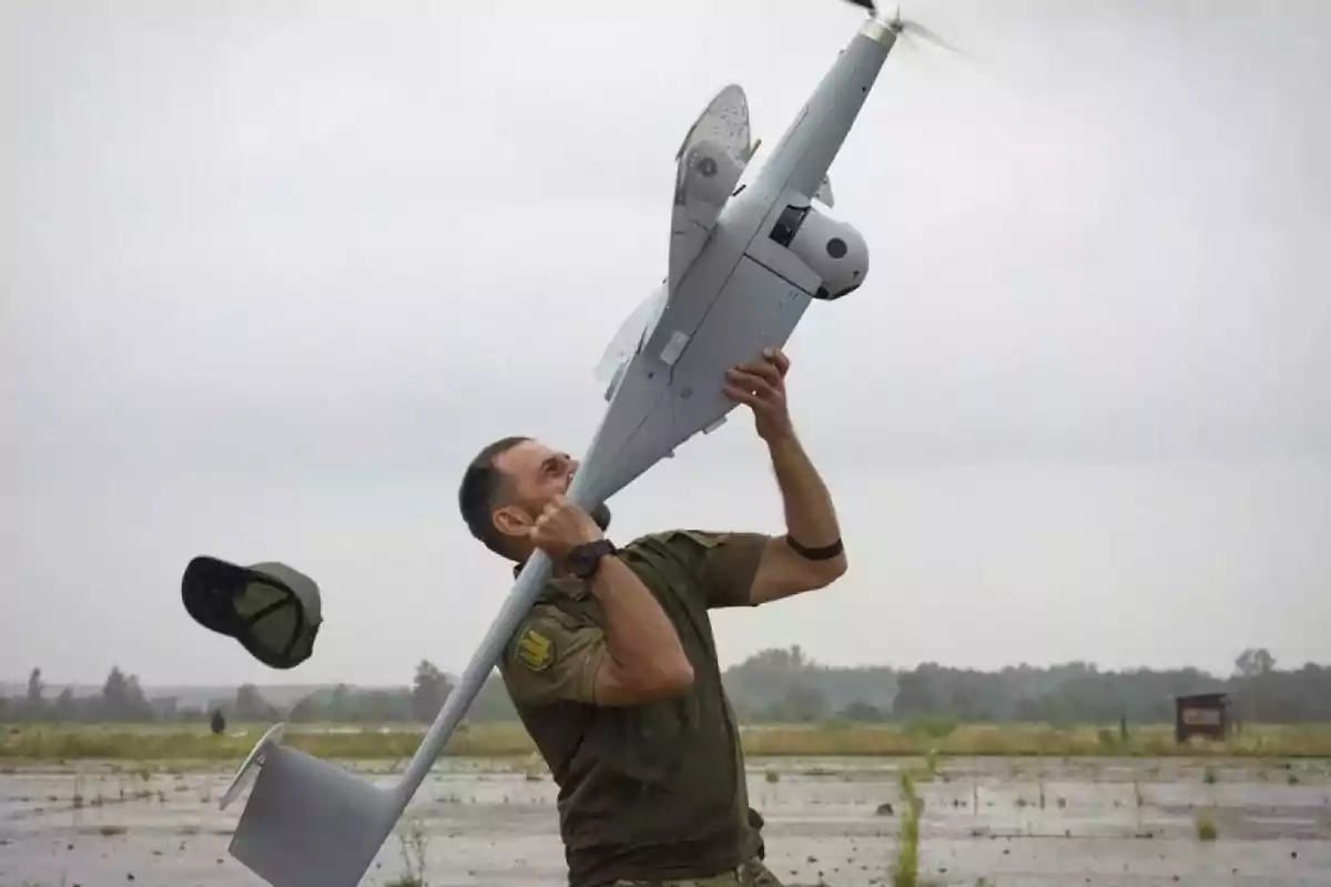 Man in military uniform launching a drone into the air in an open field while his cap flies backward Man in military uniform launching a drone into the air in an open field while his cap flies backward