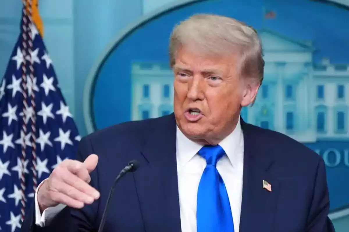 Blond-haired man in a blue suit and blue tie speaking in front of a microphone with a United States flag and a blue background behind him Blond-haired man in a blue suit and blue tie speaking in front of a microphone with a United States flag and a blue background behind him
