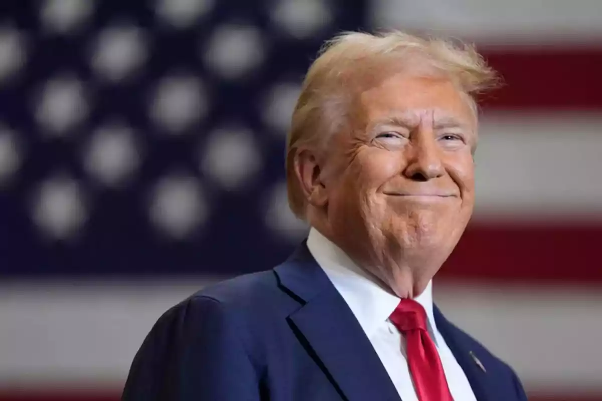 Hombre de cabello rubio con traje azul y corbata roja sonriendo frente a una bandera de Estados Unidos desenfocada