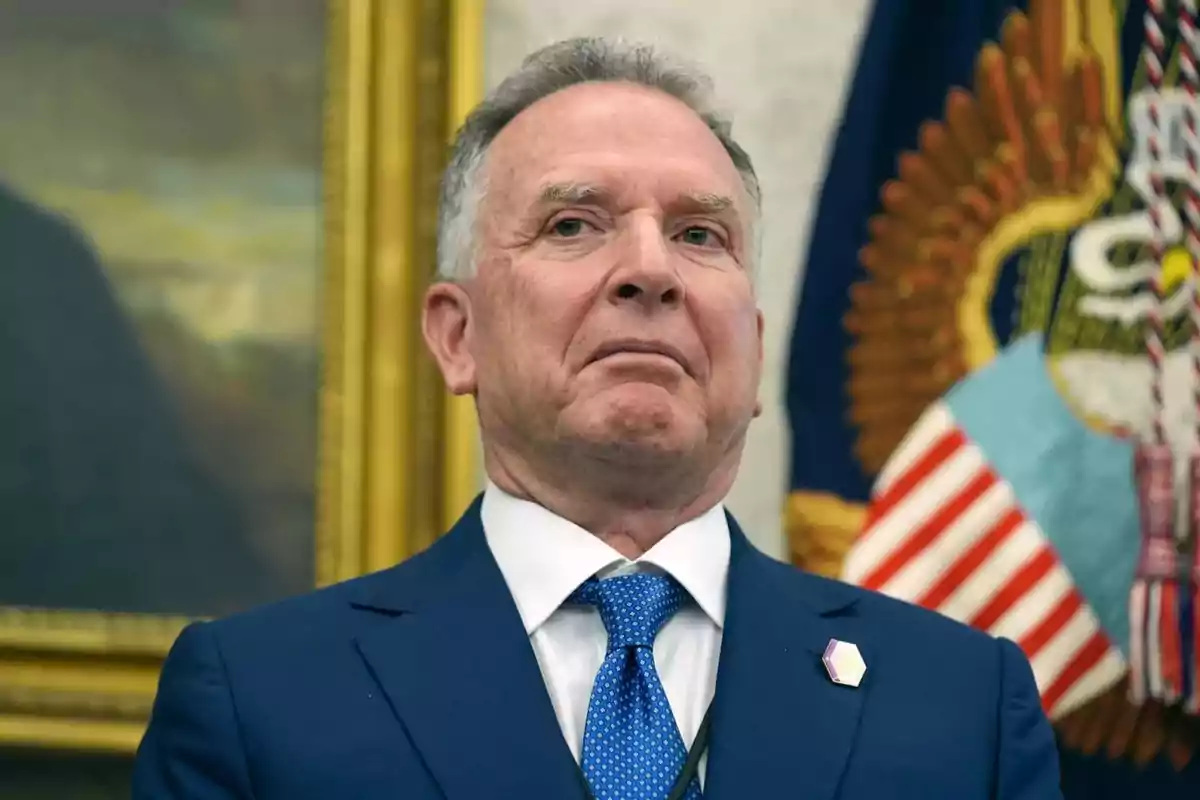 Hombre de cabello canoso con traje azul y corbata azul posando frente a una bandera de Estados Unidos y un escudo oficial Hombre de cabello canoso con traje azul y corbata azul posando frente a una bandera de Estados Unidos y un escudo oficial