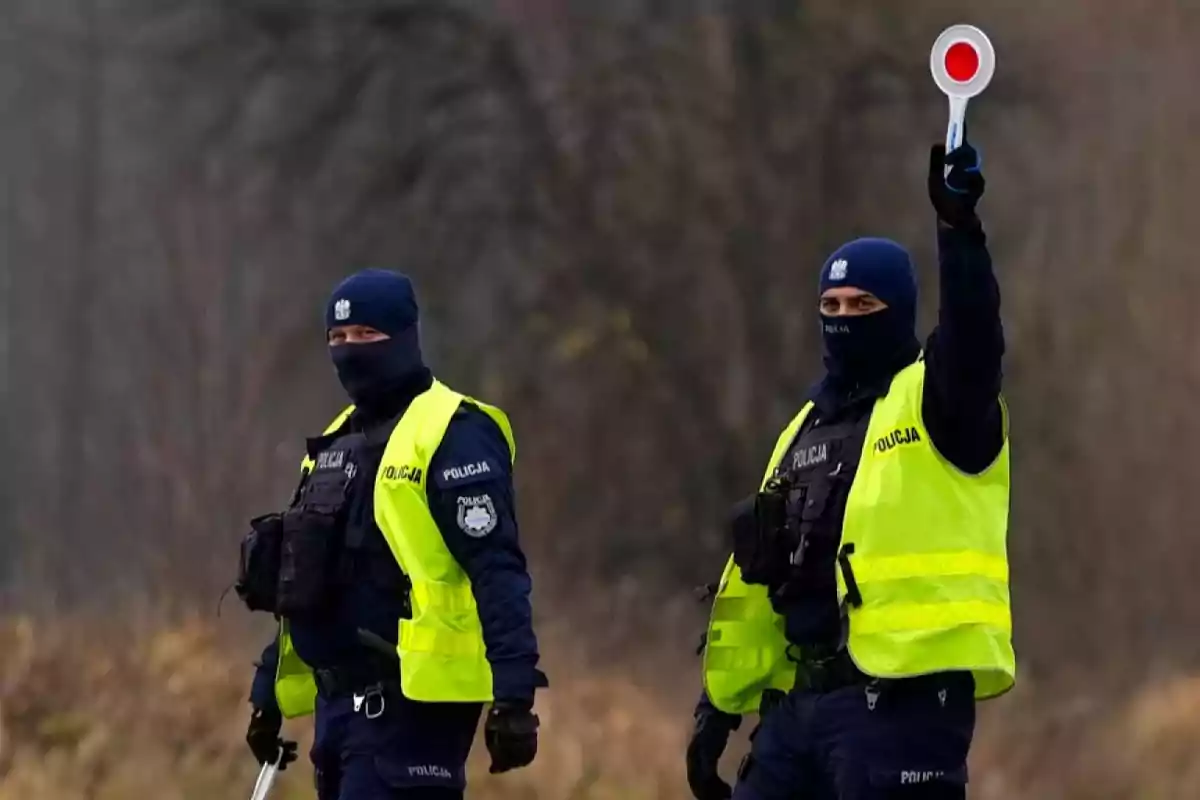 Dos policías con chalecos reflectantes y pasamontañas están de pie al aire libre, uno de ellos sostiene una señal de alto con un círculo rojo Dos policías con chalecos reflectantes y pasamontañas están de pie al aire libre, uno de ellos sostiene una señal de alto con un círculo rojo